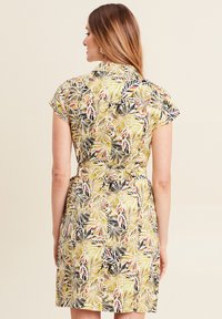 Kurzärmliges Kleid aus einem leuchtend gelben Stoff mit einem Blattmuster in Grün, Schwarz und Rot; betonter Taillenaht und Knopfkragen.