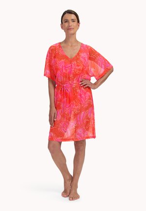 Frau steht barfuß und trägt ein knielanges Kleid in Pink und Orange mit Paisley-Muster, kurzen Ärmeln und einem Taillenband.