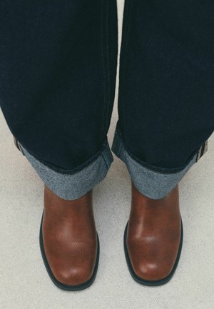 Person wearing rolled-up dark denim jeans and brown square-toe leather boots standing on a light surface.