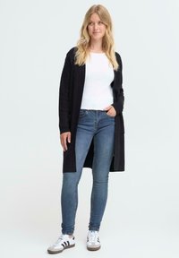 Hosszú fekete kardigán, puha anyagból, fehér bordás felső és kék szűk farmernadrág felett viselve. Fehér sportcipővel kiegészítve, fekete csíkokkal.