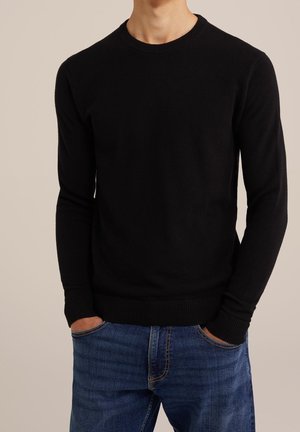 Mann trägt einen schwarzen, strukturierten Pullover mit langen Ärmeln, gerippten Bündchen und Saum, kombiniert mit blauen Jeans, Hände in den Taschen.