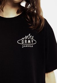 Czarna bawełniana koszulka z krótkim rękawem z białym haftowanym logo "GRMY" z motywem płomieni. Prosty design, okrągły dekolt, miękka struktura.