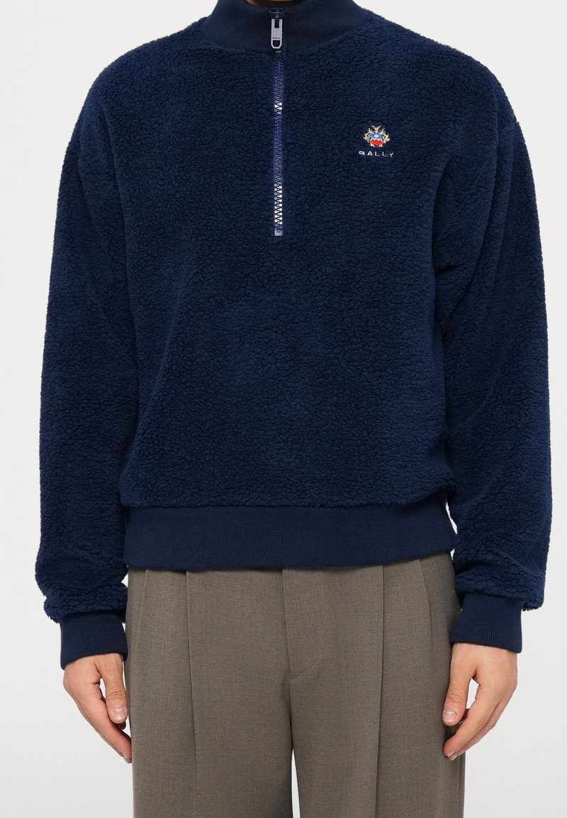 Personne portant un pull polaire bleu marine avec une fermeture éclair quart de zip et un petit logo brodé, associé à un pantalon plissé gris.
