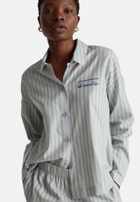 Camisa abotonada con rayas en verde y lavanda, con un logo y puños. Tela suave con un ajuste relajado y botones visibles.