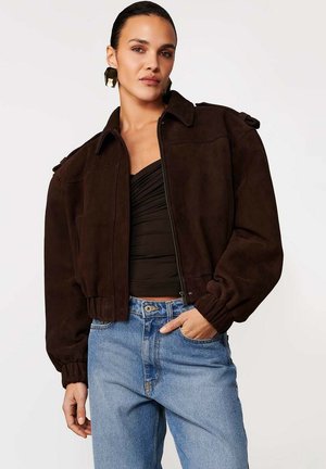 Kobieta w ciemnobrązowej krótkiej zamszowej kurtce na czarnym marszczonym topie i jasnoniebieskich dżinsach z wysokim stanem, z jedną ręką w kieszeni.