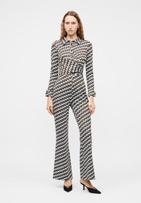 Kobieta w wzorzystym kombinezonie z długimi rękawami i rozszerzanymi nogawkami, w czarnych butach na obcasie, stojąca na jednolitym białym tle.