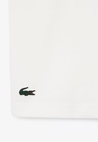 Camiseta de algodón blanca con un pequeño logo de cocodrilo verde y boca roja en el dobladillo. Textura suave, diseño simple, de manga corta.