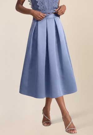 Frau trägt einen knielangen, plissierten hellblauen Rock, Spitzenoberteil und silberne offene High-Heel-Sandalen, steht vor einem beigen Hintergrund.