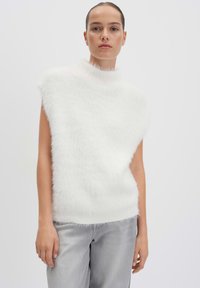 Weißes ärmelloses Oberteil mit hohem, flauschigem Kragen und weicher Textur. Kombiniert mit hellgrauen, locker sitzenden Jeans. Einfaches und klares Design.