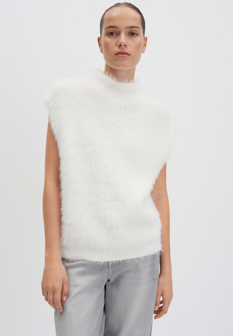 Weißes ärmelloses Oberteil mit hohem, flauschigem Kragen und weicher Textur. Kombiniert mit hellgrauen, locker sitzenden Jeans. Einfaches und klares Design.