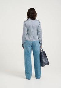 Lys grå satinbluse med lange ærmer, parret med højtaljede blå denim bukser med vidde. Modellen holder en sort læder tote taske.