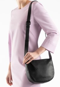 Black leather crossbody bag with a curved shape, adjustable strap, flat front pocket detail, and subtle branding on the bottom.