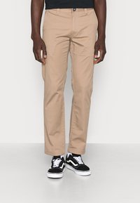 Pantalon en coton couleur fauve à coupe droite avec passants pour ceinture, associé à des baskets noires et blanches à lacets.