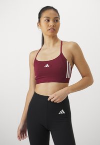 Bordeaux sportcrop-top med vit Adidas-logga och tre vita ränder på ena sidan, matchas med svarta högmidjade leggings. Slät textur.