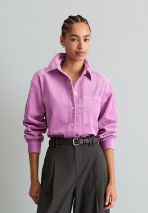 Donna con capelli intrecciati che indossa una camicia rosa di velluto a coste abbottonata infilata in pantaloni grigio scuro a vita alta con cintura nera.