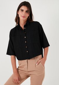 Black short-sleeve button-up shirt with vertical stripes, featuring two chest pockets and wooden buttons. Worn with beige pants.