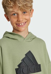 Felpa verde con un grande logo nero di Adidas sul davanti. Il tessuto sembra morbido; presenta un cappuccio con cordino e una vestibilità rilassata.