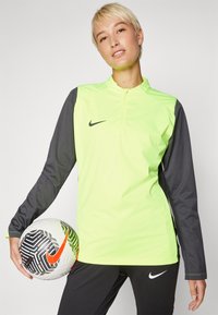 Dugi rukav sportski top s polu-zip ovratnikom, svijetlo neon žute prednje strane i tamnosivim rukavima, s Nike logotipom na prsima.