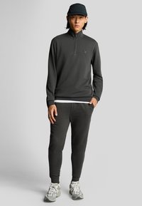 Felpa zip grigia scura con polsi a coste, abbinata a pantaloni jogger coordinati. Indossata con un berretto nero e scarpe da ginnastica grigie chiare.