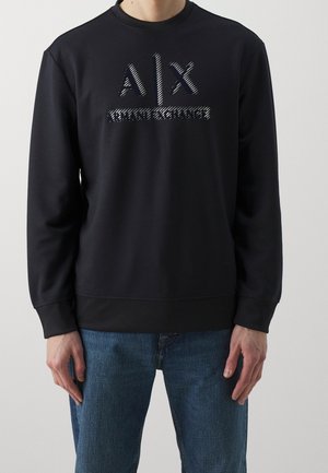 Osoba ubrana w czarną bluzę z długim rękawem z białym logo "A|X Armani Exchange", w połączeniu z niebieskimi dżinsami, stojąca na jednolitym tle.