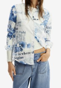 Langärmliges Hemd mit Knöpfen, das ein blaues Blumen- und Textmuster auf weißem Stoff zeigt. Das Hemd hat einen lockeren Schnitt und einen leicht geschwungenen Saum.