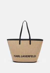 Béžová taška z rákosu s černými koženými doplňky a rukojeťmi. Má široký otevřený design a výrazné logo "KARL LAGERFELD" na přední straně.