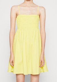 Gul linneklänning med smockad bodice och utsvängd kjol. Tillverkad av lättviktsmaterial, den har tunna axelremmar och når till mitten av låret.