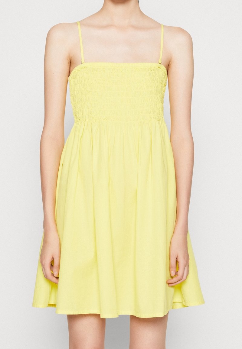 Gul linneklänning med smockad bodice och utsvängd kjol. Tillverkad av lättviktsmaterial, den har tunna axelremmar och når till mitten av låret.