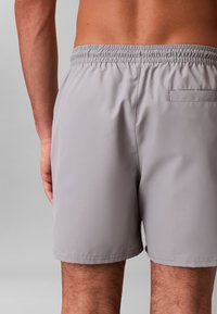 Mężczyzna ubrany w jasnoszare szorty kąpielowe z elastycznym pasem i tylną kieszenią, widoczny od tyłu, na jednolitym szarym tle.