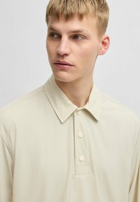 Hellbeige Poloshirt mit spitzem Kragen, verbreitertem Knopfverschluss und glatter Stoffstruktur mit dezenten Stitching-Details.