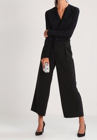 Chemise noire à manches longues avec un col en V, associée à un pantalon noir ample. Accessoirisée d'une pochette argentée texturée et de talons hauts noirs.