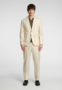 Beżowy dwuczęściowy garnitur z jednorzędową marynarką, klapami w kształcie notcha i dopasowanymi spodniami; połączony z ciemnozielonym polo i czarnymi butami.