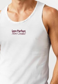 White cotton tank top featuring a rounded neckline and printed text in burgundy: "Less Perfect More Creative." Simple design, no visible patterns.