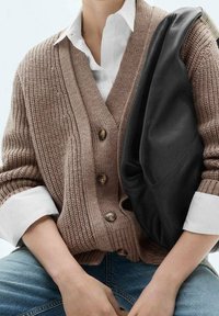 Hnědý pletený cardigan s dřevěnými knoflíky přes bílé tričko, spárovaný s modrými džíny. Na rameni odpočívá černá kožená kabelka.