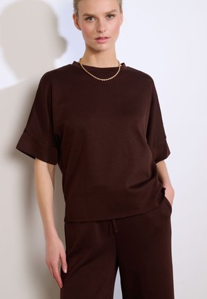 Mujer con top de manga corta marrón oscuro y pantalones a juego, collar de cadena dorada, de pie contra una pared blanca con una mano en el bolsillo.