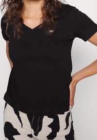 Czarna bawełniana koszulka z dekoltem w serek, krótkimi rękawami i małym logo na piersi. Zestawiona z wzorzystymi spodniami w odcieniu beżowym i czarnym.