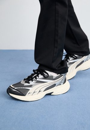 Personne portant un pantalon noir et des baskets de sport argentées, noires et blanches se tenant sur un tapis bleu.