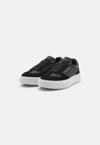 Svarte sneakers laget av semsket skinn og lær, med rund tå, flat hvit gummisåle og svarte snørebånd med metallhten.