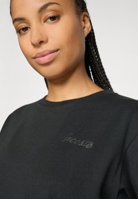 Moteris su pintomis plaukais, vilkinti juodą Lacoste sportinį megztinį, šiek tiek šypsosi neutraliame šviesiai pilkame fone.