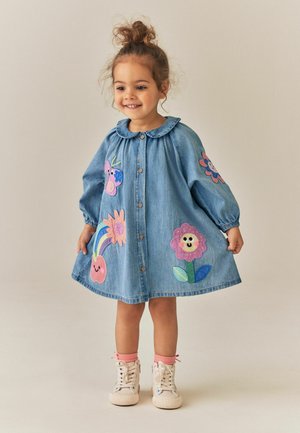 Niña pequeña sonriente vestida con un vestido vaquero azul con parches de animales y flores de dibujos animados coloridos y zapatillas blancas con calcetines rosas.