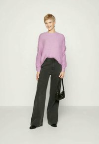 Νέος άνθρωπος με κοντά ξανθά μαλλιά, φορώντας ένα λιλά πουλόβερ, φαρδιά μαύρα παντελόνια, μαύρα παπούτσια και κρατώντας μία μικρή μαύρη τσάντα.