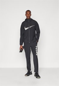Nike sort vindjakke med et reflekterende logo, parret med sorte bukser med hvide mønsterdetaljer og sorte sportsko.