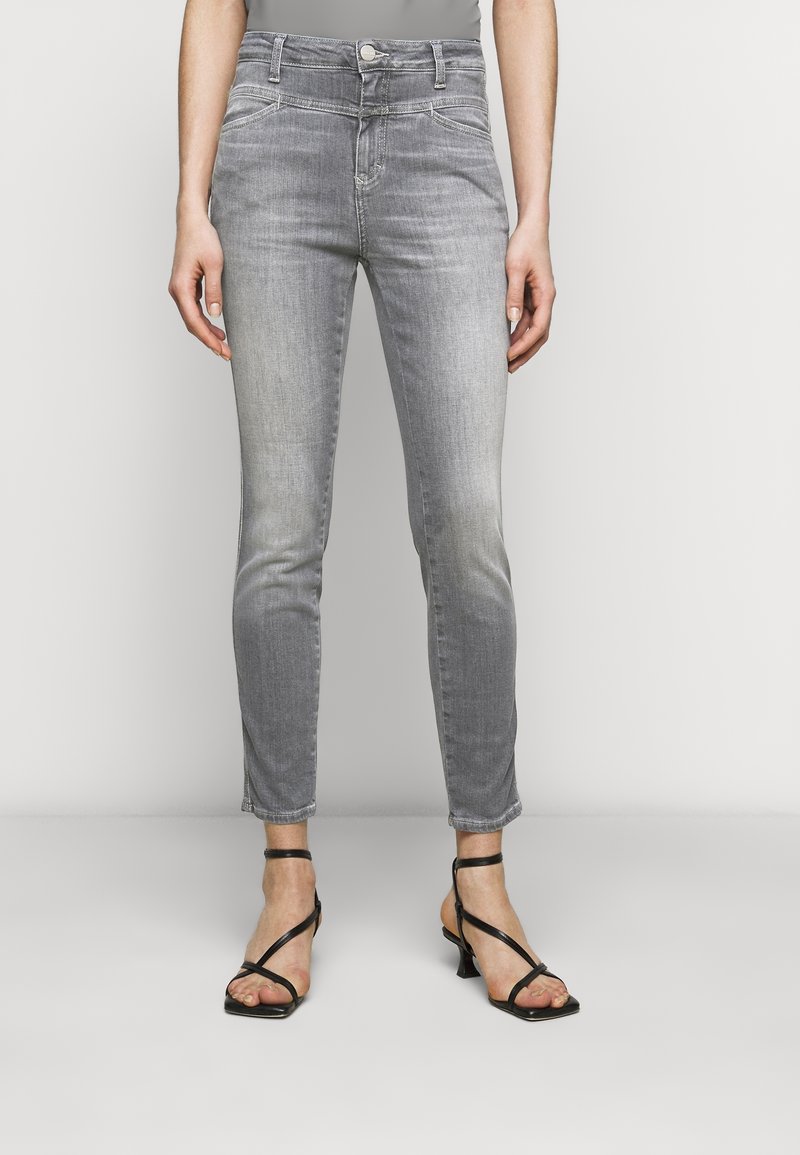 Vaqueros de mezclilla gris con un ajuste Slim, cintura de altura media, costuras en contraste y un diseño de cinco bolsillos. Combinados con sandalias de tacón negro con tiras.