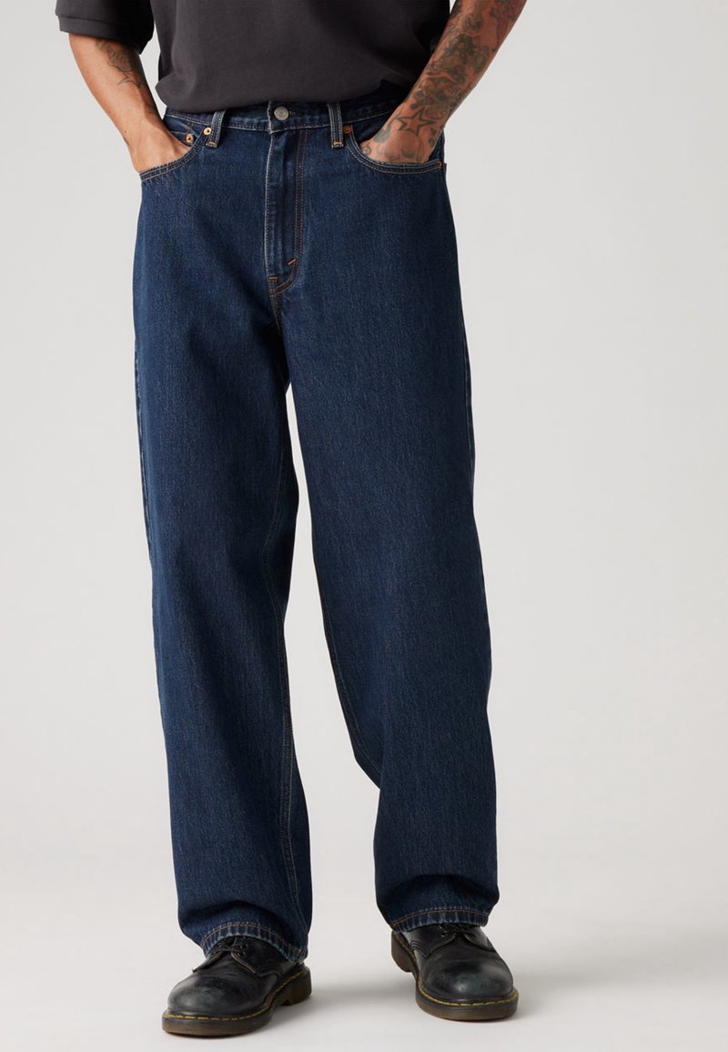Personne portant un jean large bleu foncé avec les mains dans les poches, des chaussures en cuir noires et une manche de chemise noire montrant des bras tatoués.