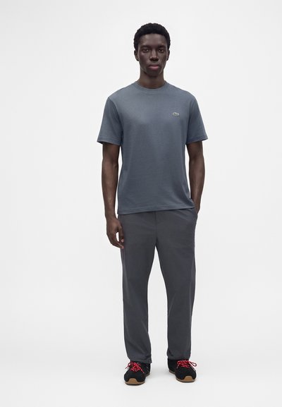 Un jeune homme est debout, portant un t-shirt gris uni, un pantalon gris foncé et des baskets noires avec des lacets rouges sur un fond blanc.