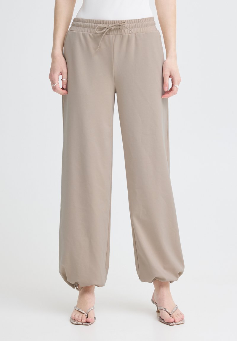 Pantalon beige à jambes larges avec taille élastique et cordon de serrage, poignets élastiques froncés, porté avec un haut blanc et des sandales à talons.
