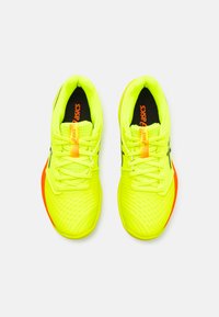 Scarpe da ginnastica giallo brillante con una tomaia in rete testurizzata, accenti neri del logo e una suola arancione. Progettate per le prestazioni e il comfort.
