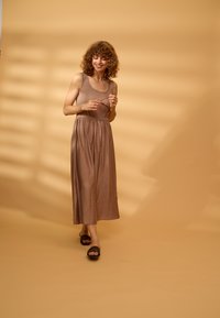 Femme aux cheveux bouclés portant une robe marron sans manches et des sandales noires, tenant des lunettes de soleil, marchant avec un doux sourire sur un fond beige.