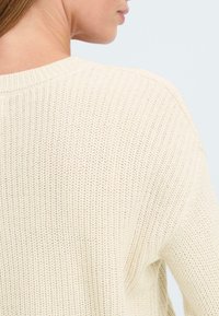 Maglione a maglia color crema con una struttura testurizzata, girocollo e coste visibili lungo il colletto e le cuciture delle spalle.