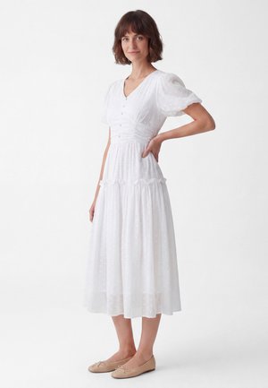 Woman with short brown hair wearing a white midi dress with puff sleeves and beige ballet flats, standing with one hand on her hip.